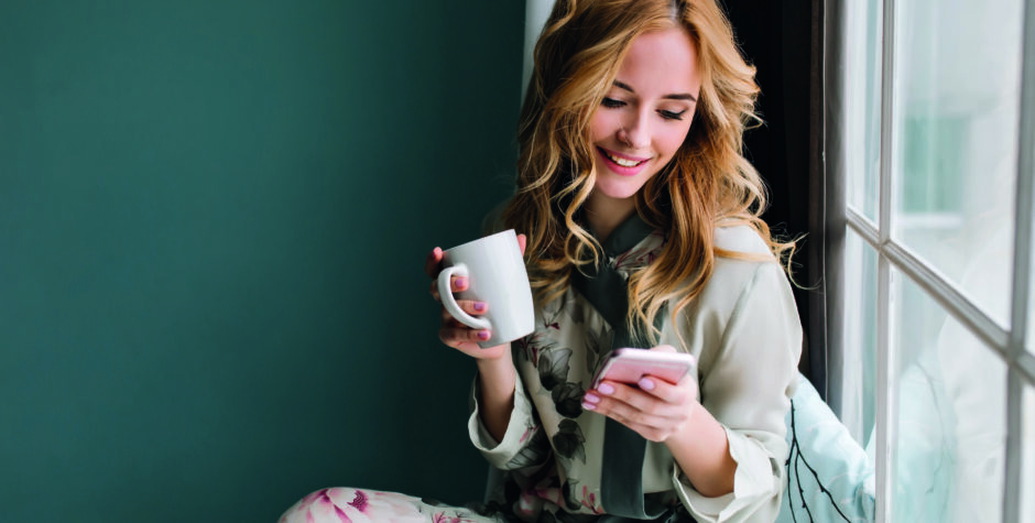 Pretty blonde girl sitting on window sill with cup of coffee, tea and smartphone in hands. She has long blonde wavy hair, smile and looking at her phone. Wearing beautiful silk pajama.